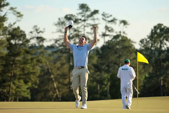 Rory McIlroy leaps in the air with both arms raised beside the 18th green